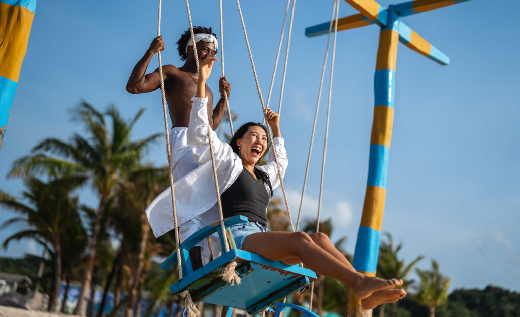 Happy interracial couple enjoying a fun beach day, swinging under palm trees on a tropical vacation